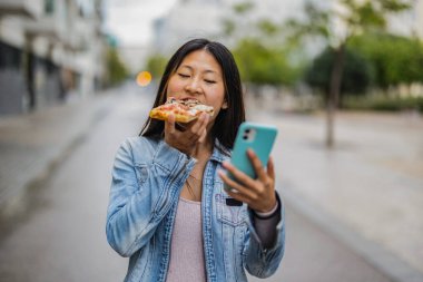 Sokaktaki Asyalı kadın pizza yerken sosyal medyayı akıllı telefonuyla kontrol ediyor.