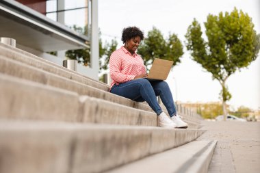 Genç, kıvrımlı Afrikalı iş kadını öğrenci şehirde merdivenlerde dinleniyor. Bilgisayarlı ekran izleme videosuna bakıyorum. İş yerindeki bitirilmiş iş üzerinde dinleniyor..