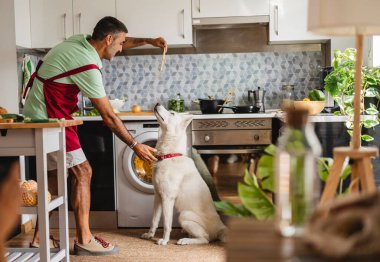 Hispanic mature man cooking with dog in kitchen feeds spaghetti