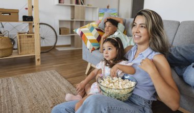 Güzel, genç İspanyol anne ve çocukları televizyon izliyor, patlamış mısır yiyor ve evde kanepede otururken gülümsüyorlar.