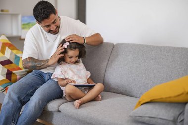 İspanyol baba küçük kızının saçını kanepede tarıyor.