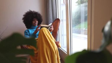 Afrikalı Amerikalı bir kadın kış günü evinde kanepede oturmuş kitap okuyor.