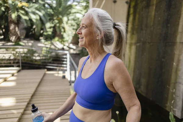 Senior woman smiles during energy drink break in outdoor gym - Mature gray-haired woman taking care of her health by doing sports -