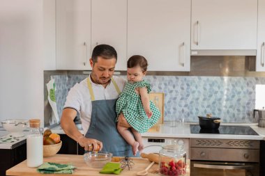 Bekar Latin Amerikalı baba, bebeğini kucağında evde pişiriyor.