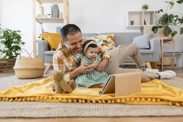 Latin American father and baby daughter using digital tablet, watching cartoons in living room at home
