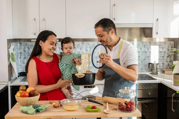 dad and mom cooking at home with baby in arms - beautiful Latin American family preparing delicious food in the kitchen -