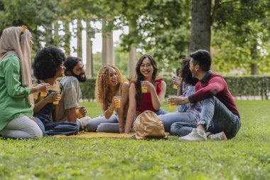 Parktaki bir grup çok ırklı arkadaş birlikte eğleniyor ve içiyorlar.