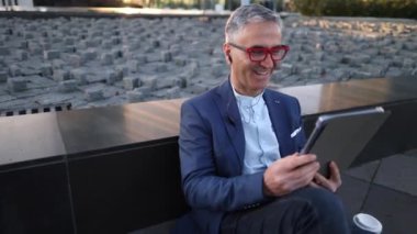 Şehir arka planında tablet kullanan gözlüklü ve takım elbiseli olgun bir erkek.