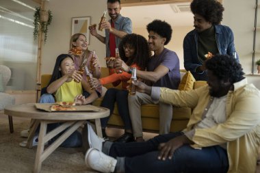 Çok ırklı arkadaşlardan oluşan mutlu bir grup, akşam yemeğinde pizza yerken dairenin oturma odasında kadeh kaldırıyor.