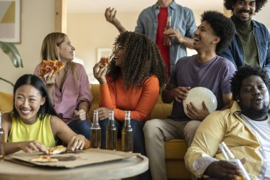 Bir grup çok ırklı yetişkin arkadaş kanepede oturmuş pizza yiyor ve üniversiteden sonra arkadaşlıklarını kutluyorlar.