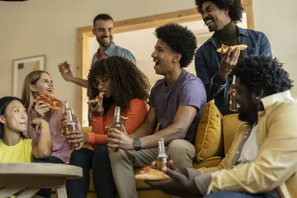 Group of multiethnic friends on a couch eating pizza and drinking beer at a house party, while laughing telling stories
