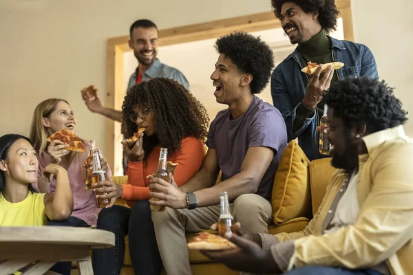 Group of multiethnic friends on a couch eating pizza and drinking beer at a house party, while laughing telling stories