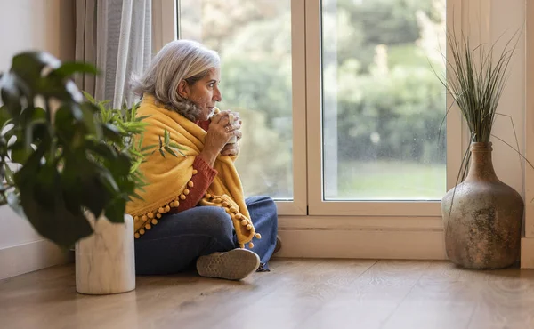 senior woman sitting on the floor of her apartment drinking hot cup drink next to the window covered