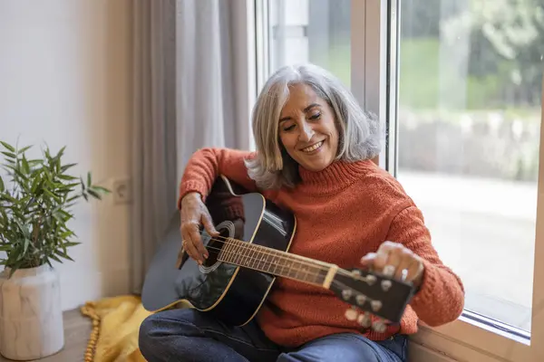 Senior woman tuning her guitar sitting on the couch in her living room