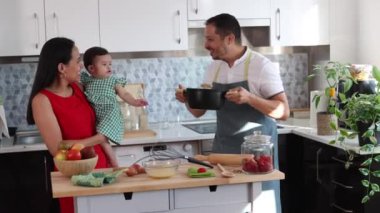 Ebeveynler arasındaki işbirliği. Kocası yemek pişirirken, karısı da kollarında bebekle ona eşlik ediyor.