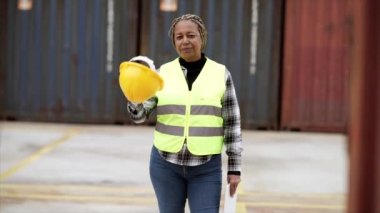 Güvenlik yeleği giymiş Afro-Amerikan kadın endüstriyel mühendisin lojistik merkezinde çalışan kaskını takarken gülümseyen portresi. Konteynır Terminal Yöneticisi.
