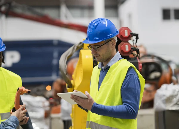 Modern factory: Male engineer project manager standing wearing safety jackets, helmets, working in workshop, talking, using digital tablet and monitoring CNC machinery assembly line