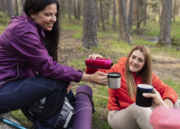 group of multicultural female friends resting in the forest while having a coffee with their thermos and sharing time together laughing