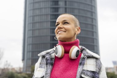 Arkaplanda gökdelenler ve boynunda kulaklıklarla gökyüzüne bakan traşlı genç kadın portresi