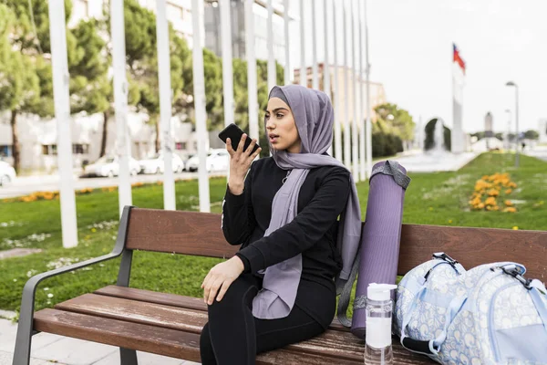 veiled woman recording audio with her cell phone while waiting for her gym partner sitting on a bench