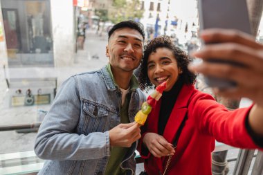 Çok kültürlü bir çift şehirde birlikte vakit geçirirken akıllı telefonlarıyla selfie çekiyor.