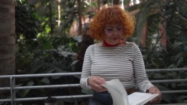 Kızıl saçlı, gözlüklü olgun bir kadın. İstasyon bahçesinde oturmuş kitap okuyor.
