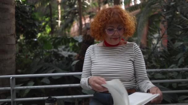红头发的成熟女人 戴着眼镜 坐在车站花园 — 图库视频影像