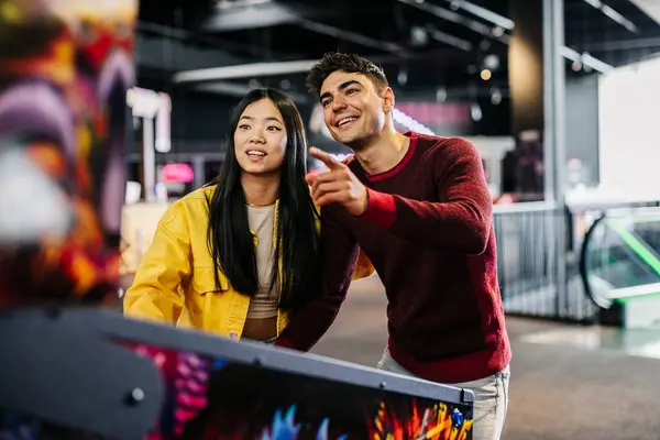 generation z multiracial couple playing pinball machine in the game room