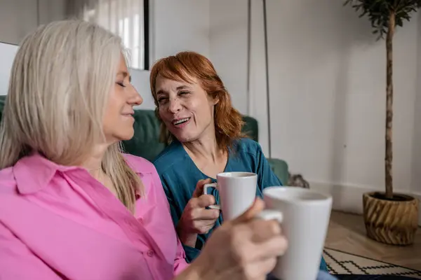 Two joyful mature friends share a laugh over coffee at home.