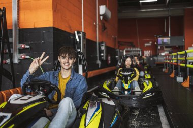 Gülümseyen genç adam, arka planda odaklanmış bir kadın sürücüyle go-kart yarışına başlamadan önce barış işareti veriyor..