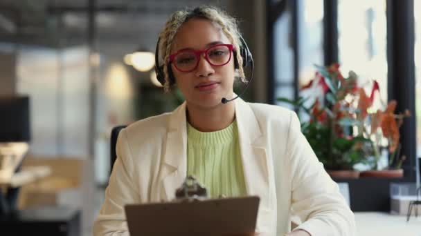 可接近的客户服务代表 带着耳机微笑坐在她的办公桌前 — 图库视频影像