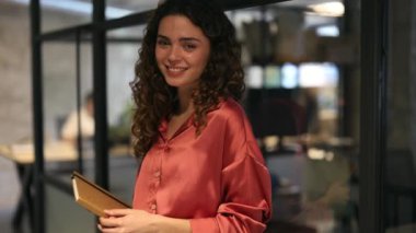 Modern bir ofis koridorunda elinde kitap tutan ve içten gülümseyen neşeli genç bir kadın..