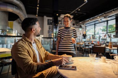 Çizgili tişörtlü arkadaş canlısı garson, çağdaş bir kafede oturan bir erkeğe kahve ikram ediyor..