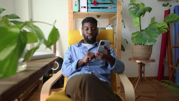 陽気なアフリカ系アメリカ人男性が自宅の椅子でリラックスし ヘッドフォンで音楽を聴き スマートフォンを保持しています 自宅でスマートフォンでヘッドフォンで音楽を楽しむ楽しい男 — ストック動画