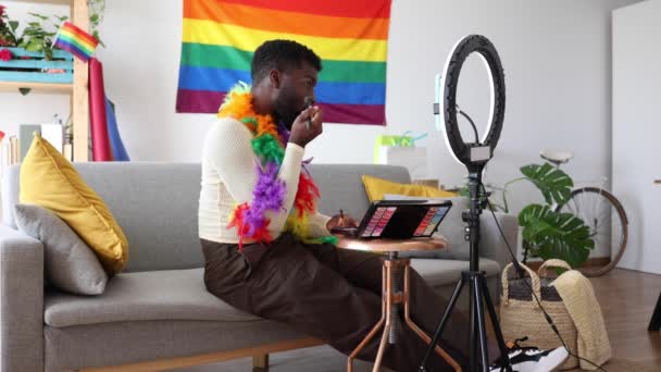 カラフルなボロで飾られた男性は リングライトの前に化粧を施し プライドをテーマにしたビューティーチュートリアルを鮮やかな外観にしています Pride Themedチュートリアルのためのメイクを適用するコンテンツクリエイター — ストック動画