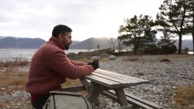 Rahat yün ceketli bir adam, bir piknik masasında huzur dolu, çakıl taşlı bir manzaranın ortasında yalnızlıktan zevk alır..