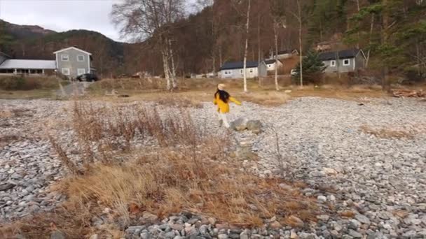 一位身穿鲜艳的黄色上衣 头戴贝尼帽的妇女欢快地走过一块石质的海岸 远处是斯堪的纳维亚人的家 — 图库视频影像