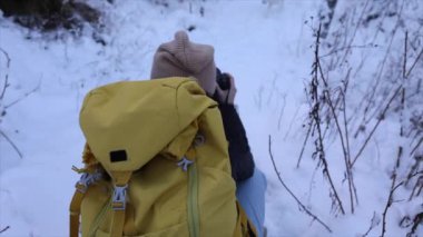 Sarı sırt çantalı ve bej bereli genç bir kadın fotoğrafçı kamerasıyla kış güzelliğini yakalıyor. Karlı Manzara 'da Genç Kadın Fotoğrafçı