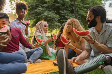 Bir grup arkadaş kahkaha ve karpuzu paylaşıyor, bereketli bir parkta yazın tadını çıkarıyorlar..