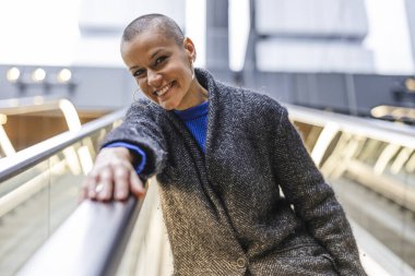 Paltolu ışıl ışıl olgun bir kadın modern bir ortamda yürüyen merdivene binerken kendine güveni tamdır..