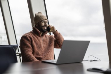 Bereli neşeli bir genç adam iş yerindeki bilgisayarının yanında telefonuyla hoş bir sohbet ediyor..