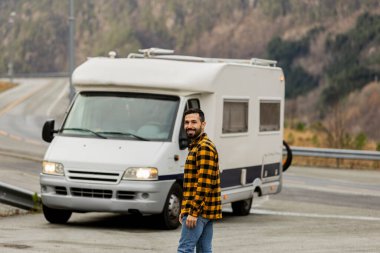 Joyful traveler with a beard stands by his camper van on a scenic mountain road, ready for adventure. clipart