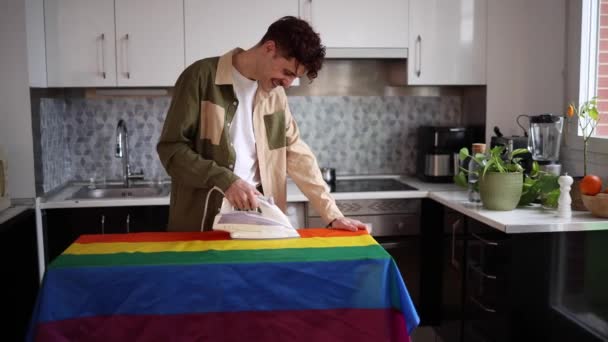 一个男人在熨斗板上小心地熨烫着鲜艳的彩虹旗 为他在当代厨房里做的家务活感到自豪 — 图库视频影像