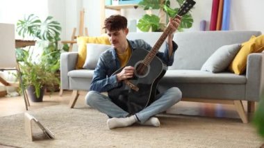 Rahatlamış genç bir yetişkin, akustik gitarını ayarlar, huzurlu evinde uyumlu bir atmosfer yaratır..
