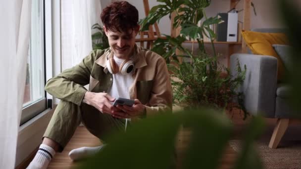Sentado Junto Ventana Joven Involucra Con Teléfono Inteligente Auriculares Listos — Vídeos de Stock