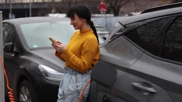 一位穿着生机勃勃的芥末毛衣的心满意足的女人在充电站插入的电动汽车旁边用她的智能手机 电动汽车收费时妇女检查电话 — 图库视频影像
