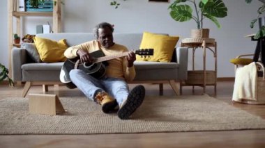 Yaşlı bir beyefendi, neşeli bir ifadeyle akustik gitar çalar, evde halı döşeli bir zeminde gelişigüzel oturur.
