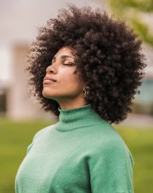 Afro saç stili nefes kesen kendine güvenen genç bir kadın, canlı yeşil bir süveter giymiş, sakin bir açık hava ortamında derin derin düşünüyor..