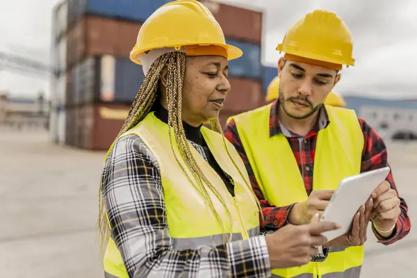 stock image Two construction workers, a middle-aged African American woman and a young Hispanic man, using a tablet in a shipping yard.