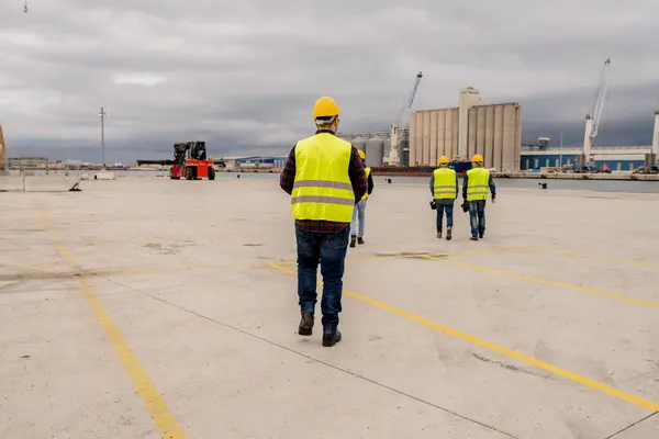 stock image A male construction worker leads a group of colleagues across a spacious port, emphasizing leadership and teamwork in an industrial setting.